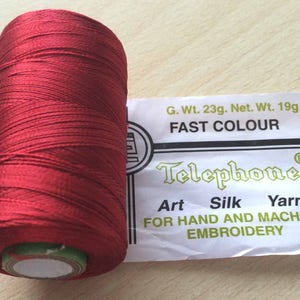 Può includere: Un'bobina di filo di seta artificiale rosso, etichettata "Telephone Art Silk Yarn" e "Fast Colour". L'etichetta indica anche "Per ricamo a mano e a macchina". La bobina pesa 23 grammi lordi e 19 grammi netti.