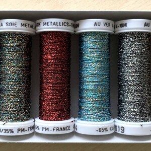 Puede incluir: Seis carretes de hilo metálico en una variedad de colores, incluyendo azul, verde, rojo, turquesa, plata y oro. Los carretes están etiquetados con el texto "AU VER A SOIE METAL DIE METALLICS-1", "AU VER-10M", "AU", "AU VER A", "0 -65%", "CU/35% PM-FRAO% PM-FRANCE", "4 -65% CU 019", y "-65% CU".