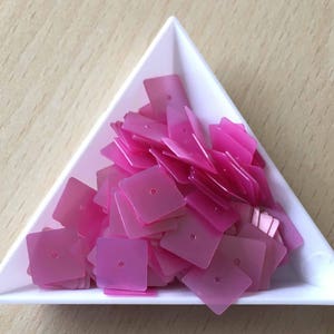 May include: A white triangular tray filled with pink iridescent sequins. The sequins are square shaped with a small hole in the centre.