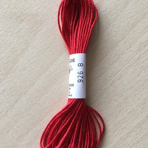 Può includere: Una bobina di filo da ricamo rosso brillante. L'etichetta recita "926 B" su una fascia bianca. Il filo è ben avvolto e appare morbido e setoso. Lo sfondo è un legno chiaro.