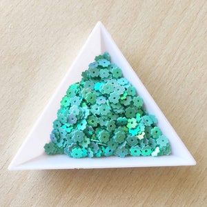 May include: A white triangular dish filled with teal iridescent flower-shaped sequins.