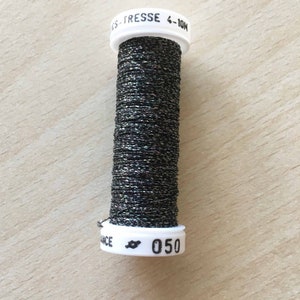 Puede incluir: Una bobina de hilo metálico negro con purpurina plateada. La bobina está etiquetada con "CS - TRESS 4-10H" y "050".