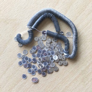 Könnte beinhalten: Ein Haufen silberner Pailletten mit gewelltem Rand, neben einer Rolle silbernen und grauen Metallic-Zierbands.