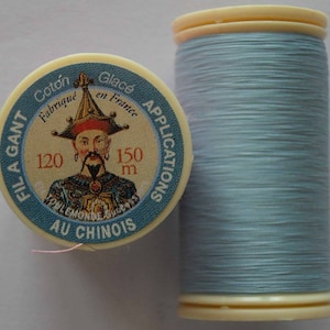 Może przedstawiać: Szpulka jasnoniebieskiej nici z etykietą, na której widnieje napis "Fila Gant Coton Glacé Fabriqué en France Applications Au Chinois" oraz zdjęcie mężczyzny w tradycyjnym chińskim kapeluszu. Etykieta zawiera również liczby "120" i "150 m".