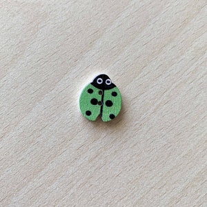 Può includere: Un bottone di legno verde a forma di coccinella con macchie nere e occhi bianchi.