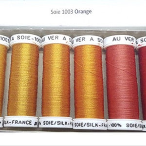 May include: A box containing six spools of Soie 1003 Orange silk thread. The spools range in colour from gold to red. The text "Soie 1003 Orange" is at the top. The brand name "Silk-France" is visible on the spool labels.