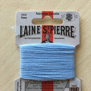 Peut inclure: Une bobine de fil Laine St. Pierre bleu clair, coloris 705, sur une étiquette blanche et rouge. L'étiquette indique "Fabrication française" et "REPRISER BRODER FESTONNER". Le fil est composé à 50% de laine et 50% de polyamide. Idéal pour la broderie et la couture.