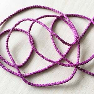 Könnte beinhalten: Nahaufnahme eines geflochtenen Bandes in Lila und Gold. Das Band ist aus einem weichen, seidenartigen Material gefertigt und hat einen dezenten Schimmer.