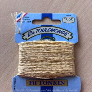 May include: A spool of Fil Tonkin thread, color 1050, made by Ets Toulemonde. The thread is a light tan color and is wrapped around a blue cardboard spool. The packaging includes the text "Fabriqué France depuis 1903" and "100% polyester."