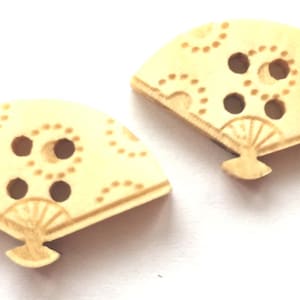 Può includere: Due bottoni in legno beige a forma di ventaglio con un motivo a pois e tre fori.