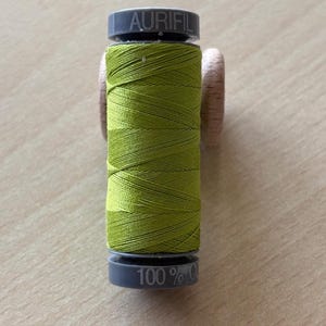 Pode incluir: Um carretel de linha Aurifil em verde vibrante. A linha é enrolada firmemente em torno de um núcleo de madeira, com a marca "AURIFIL" impressa na parte superior de plástico cinza. A parte inferior do carretel diz "100% C".
