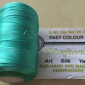 May include: A spool of teal blue art silk yarn for hand and machine embroidery. The label reads "Telephone Art Silk Yarn" and "Fast Colour". The spool weighs 23 grams gross and 19 grams net.
