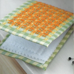 Peut inclure: Une petite couverture de livre en tissu à carreaux verts et blancs avec des broderies orange. La couverture est pliée et le tissu est doux et texturé.
