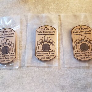May include: Three identical dog tag-shaped wooden ornaments, each individually packaged in a clear plastic sleeve. The ornaments feature the engraved text 2024 MAHS State Champions, a bowling ball and pins graphic, and a list of names including Coach Bader.