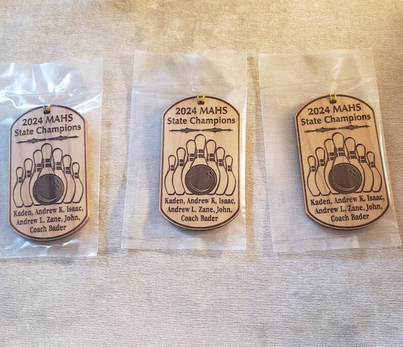 May include: Three identical dog tag-shaped wooden ornaments, each individually packaged in a clear plastic sleeve. The ornaments feature the engraved text 2024 MAHS State Champions, a bowling ball and pins graphic, and a list of names including Coach Bader.