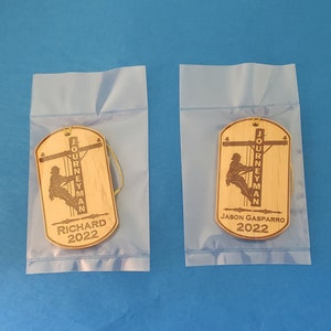 May include: Two wooden ornaments with a silhouette of a lineman working on a power line. The ornaments are in clear plastic bags. The text on the ornaments reads "Journeyman" and the names "Richard 2022" and "Jason Gasparro 2022".