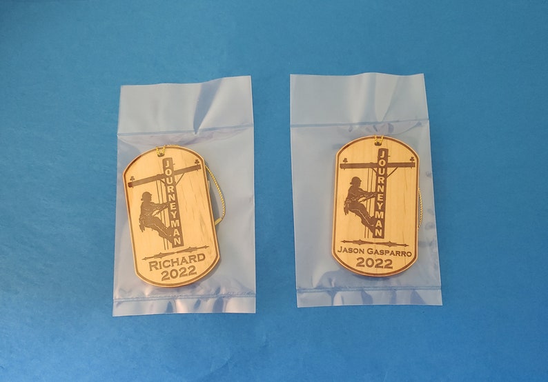 May include: Two wooden ornaments with a silhouette of a lineman working on a power line. The ornaments are in clear plastic bags. The text on the ornaments reads "Journeyman" and the names "Richard 2022" and "Jason Gasparro 2022".