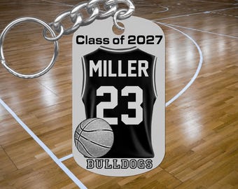 May include: A silver-toned metal keychain with a rectangular tag featuring the text Class of 2027, MILLER, 23, and BULLDOGS. The design includes a graphic of a black basketball jersey and a basketball. The keychain has a circular metal ring.