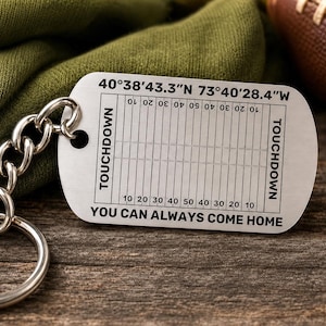 May include: A silver-toned metal dog tag keychain featuring a black engraved American football field graphic. The text reads 40°38'43.3"N 73°40'28.4"W, TOUCHDOWN, and YOU CAN ALWAYS COME HOME. The keychain includes a metal chain and a circular ring.