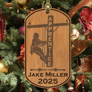May include: Wooden Christmas ornament featuring a silhouette of an electrician working on a power pole. The ornament is personalized with the text "Apprentice", "Jake Miller", and "2025".