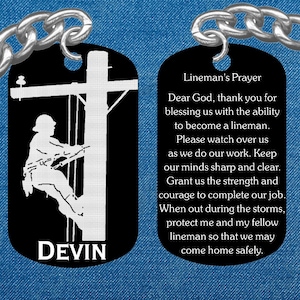 May include: Black metal keychain with a silhouette of a lineman climbing a utility pole. The other side of the keychain has a prayer for linemen. The keychain is personalized with the name "DEVIN".