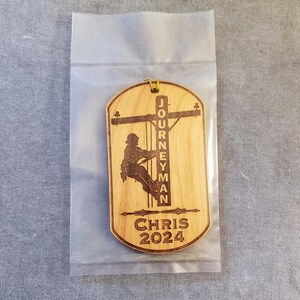 May include: A wooden ornament with a silhouette of a lineman climbing a utility pole. The ornament is engraved with the text "Journeyman Chris 2024".