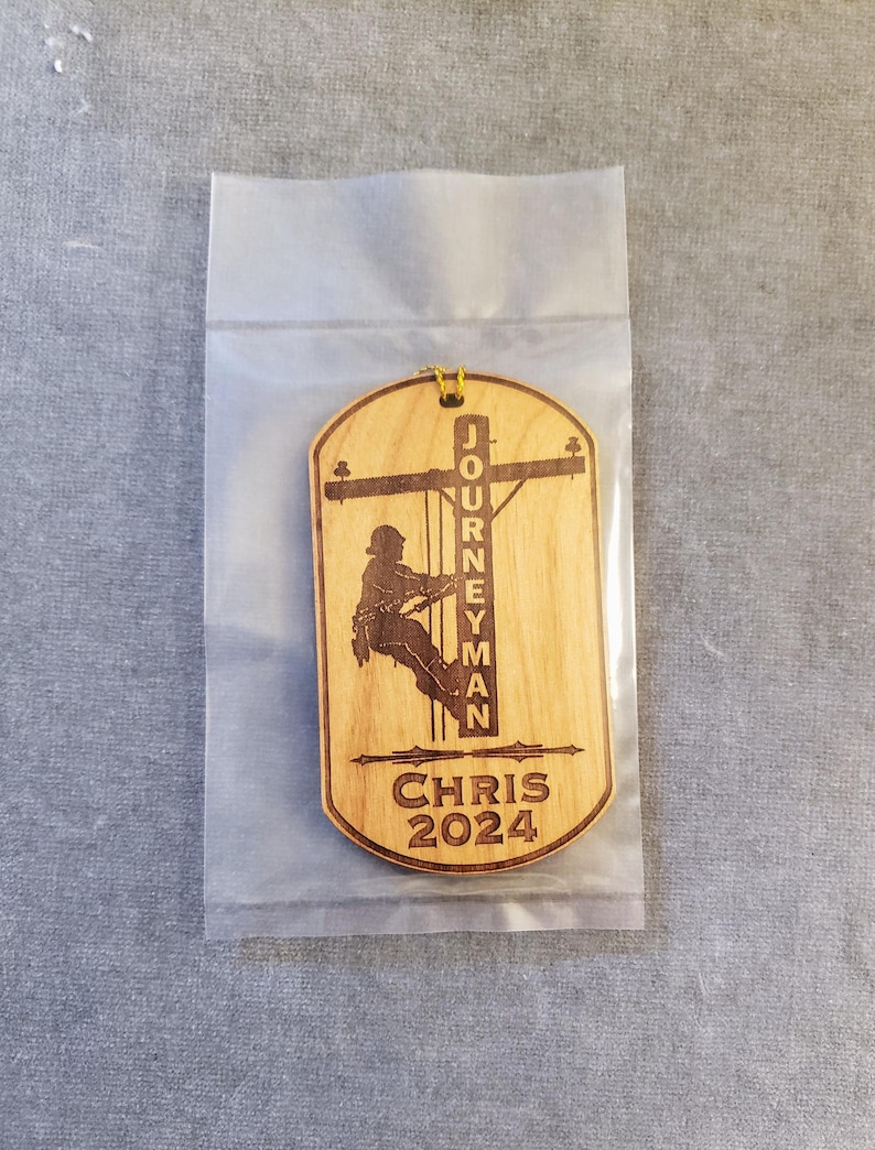 May include: A wooden ornament with a silhouette of a lineman climbing a utility pole. The ornament is engraved with the text "Journeyman Chris 2024".