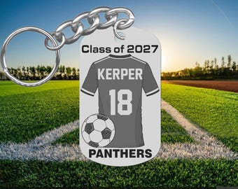 May include: A rectangular silver-toned metal keychain featuring a graphic of a gray soccer jersey with the name KERPER and the number 18 printed on it. Below the jersey is a soccer ball and the text Class of 2027 and PANTHERS.