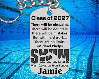 May include: A silver-toned metal dog tag keychain featuring the text Class of 2027, a quote by Michael Phelps, the word SWIM with a swimmer silhouette, West Carolton High School, and the name Jamie engraved in black lettering on the surface.