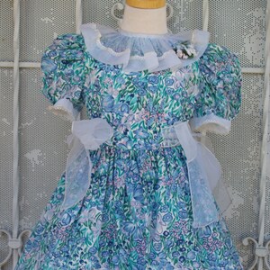 May include: A blue and white floral dress with puffed sleeves and a white lace collar. The dress has a white bow at the waist and a white tulle overlay.
