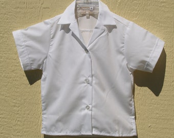May include: A white short-sleeved button-down shirt with a collar. The shirt is hanging on a silver hanger against a yellow wall.