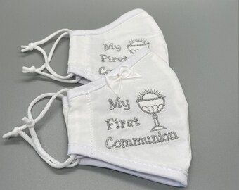 May include: Two white face masks with silver glitter embroidery. The top mask says "My First Communion" and the bottom mask says "My First Communion" with a chalice and host design.
