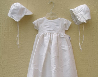 May include: A white baby christening gown with short sleeves and an embroidered detail on the bodice. Two matching bonnets with ties are displayed. The gown and bonnets are made of a textured fabric. A traditional outfit for a christening.