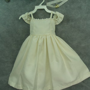 Puede incluir: Un vestido blanco sin mangas con ribete de encaje y mangas con volantes. El vestido está colgado de una percha blanca y tiene una falda fruncida.