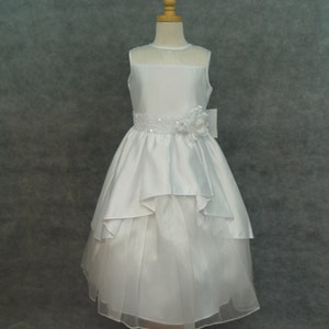 Puede incluir: Vestido blanco sin mangas con falda de capas y corpiño de satén. El vestido tiene un cinturón de cuentas y un adorno de flor blanca.