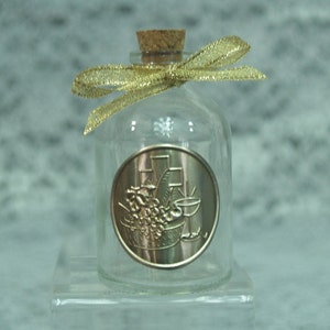 May include: A clear glass bottle with a cork stopper and a gold ribbon. The bottle has a round, gold-colored metal label with a design of grapes, a chalice, and a cross.