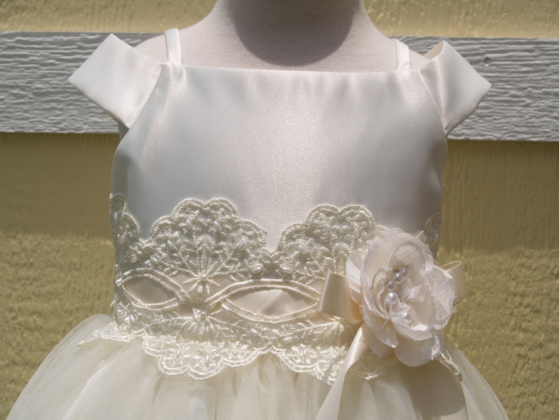May include: A white satin dress with a lace bodice and a tulle skirt. The dress has a flower embellishment on the waist.