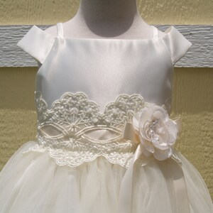 May include: A white tulle and satin dress with a lace bodice and a satin bow with a flower accent. The dress has a sleeveless design with a scoop neckline.