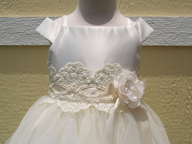 May include: A white tulle and satin dress with a lace bodice and a satin bow with a flower accent. The dress has a sleeveless design with a scoop neckline.