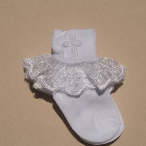 May include: White baby socks with a lace ruffle and a silver cross embroidered on the top.