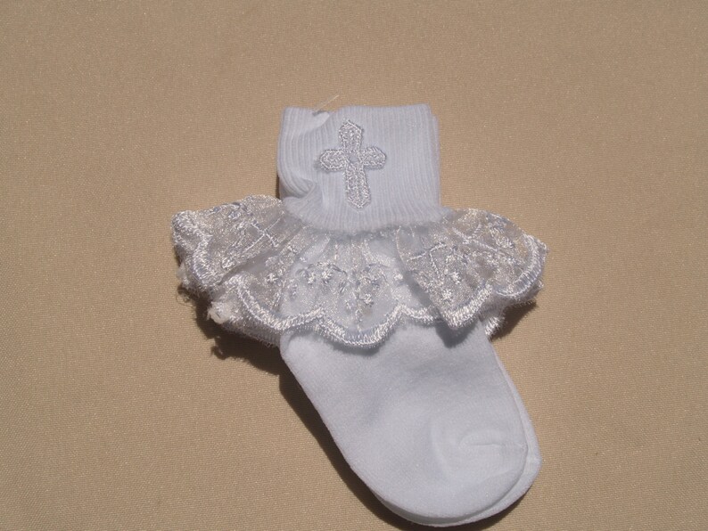 May include: White baby socks with a lace ruffle and a silver cross embroidered on the top.