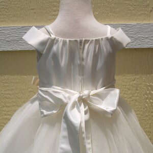 May include: White off-the-shoulder tulle dress with a satin bodice and a large satin bow at the back.