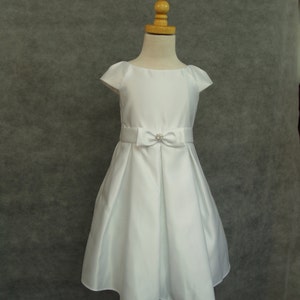 Puede incluir: Un vestido blanco de satén con un lazo y un adorno de strass en la cintura. El vestido tiene mangas cortas y una falda completa.