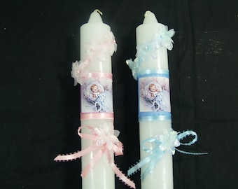 May include: Two white baptism candles with pink and blue ribbons and flowers. One candle has a pink ribbon and a picture of a baby girl, the other has a blue ribbon and a picture of a baby boy.