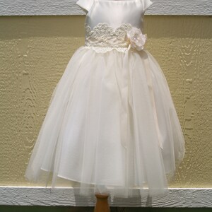 May include: A white tulle and satin dress with a lace bodice and a satin sash. The dress has a flower embellishment on the sash.