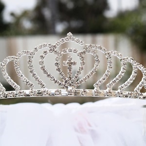 May include: A silver tiara with a cross design and rhinestones. The tiara is on a white background.