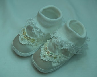 May include: A pair of baby shoes with a satin finish in a light beige color. The shoes have white lace trim and are adorned with sequins and small pearls. The ankle cuffs are white and made of a soft knit material.
