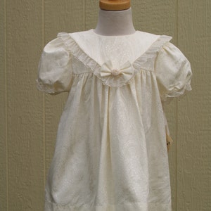 Puede incluir: Un vestido blanco con cuello de encaje y un lazo en el escote. El vestido tiene una falda fruncida y un ribete de encaje en el dobladillo.