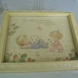 May include: A framed print with a white background and a light yellow frame. The print features a colorful illustration of two children with a lamb in a field. The text "He maketh me to lie down in green pastures... - Psalm 23:2" is printed at the top of the print.