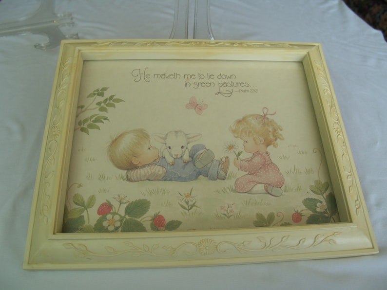 May include: A framed print with a white background and a light yellow frame. The print features a colorful illustration of two children with a lamb in a field. The text "He maketh me to lie down in green pastures... - Psalm 23:2" is printed at the top of the print.